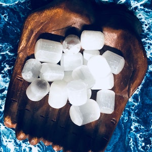 May include: A collection of white selenite tumbled stones in a wooden hand-shaped bowl. The stones are smooth and polished, with a natural, milky white color.