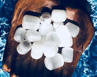 May include: A collection of white selenite tumbled stones in a wooden hand-shaped bowl. The stones are smooth and polished, with a natural, milky white color.
