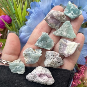 May include: A collection of eight green and pink gemstone crystals. The crystals are rough and have a variety of shapes and sizes. They are displayed on a hand.
