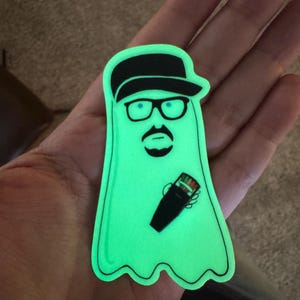 May include: A green glow-in-the-dark sticker of a ghost wearing a black hat and glasses, holding a cocktail shaker.
