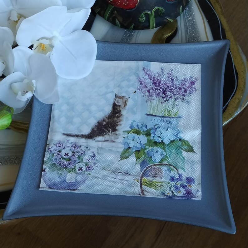 May include: A gray ceramic square tray with a white paper napkin featuring a kitten, purple flowers, and blue flowers. The napkin has the word "Flowers" printed on it.