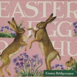 May include: A pink background with the words "Easter Egg Hunt" in white text. Two brown rabbits are jumping in the air, one facing left and the other facing right. Purple flowers are in the bottom left and right corners of the image. The artist's name, "Emma Bridgewater", is written in white text on a green banner at the bottom of the image.