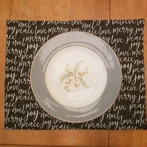 May include: A white floral patterned plate with a gray rim sits on a black placemat. The placemat is covered in white script that reads "noel", "peace", "love", "merry", and "joy". The placemat is rectangular.
