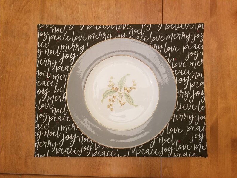May include: A white floral patterned plate with a gray rim sits on a black placemat. The placemat is covered in white script that reads "noel", "peace", "love", "merry", and "joy". The placemat is rectangular.