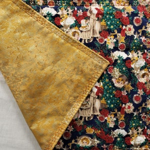 May include: A quilted Christmas table runner with a gold-colored, textured underside. The top side features a festive pattern of angels, Christmas trees, red roses, and stars on a navy blue background. The edges are stitched with gold thread.