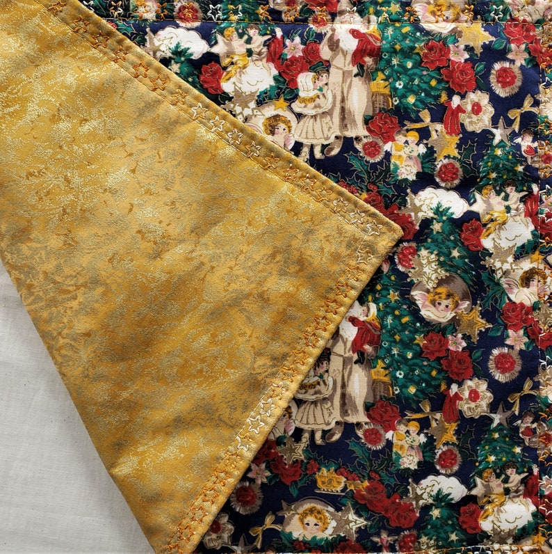 May include: A quilted Christmas table runner with a gold-colored, textured underside. The top side features a festive pattern of angels, Christmas trees, red roses, and stars on a navy blue background. The edges are stitched with gold thread.
