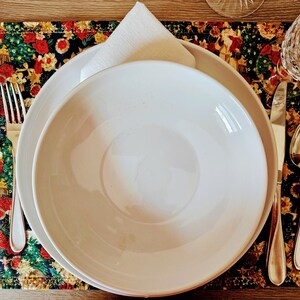 May include: A formal place setting with a white bowl and plate, a folded white napkin, and silverware. The setting is on a colorful placemat with a floral and holiday design. A glass is visible in the background.