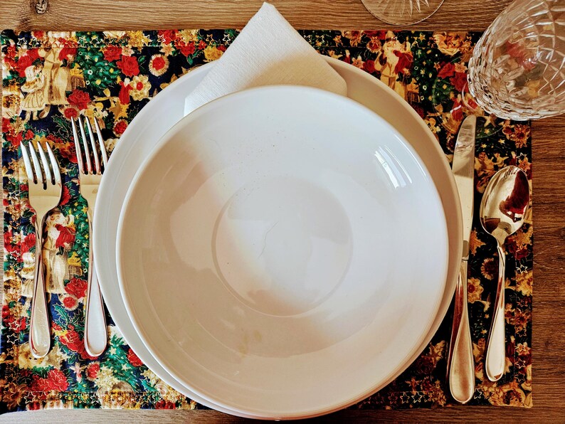 May include: A formal place setting with a white bowl and plate, a folded white napkin, and silverware. The setting is on a colorful placemat with a floral and holiday design. A glass is visible in the background.