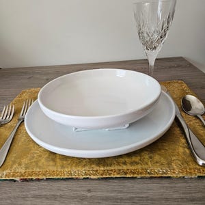 May include: A white ceramic bowl and plate set on a gold placemat, with silverware and a crystal wine glass. The bowl sits atop the plate, creating a layered effect. The setting is on a wooden table.