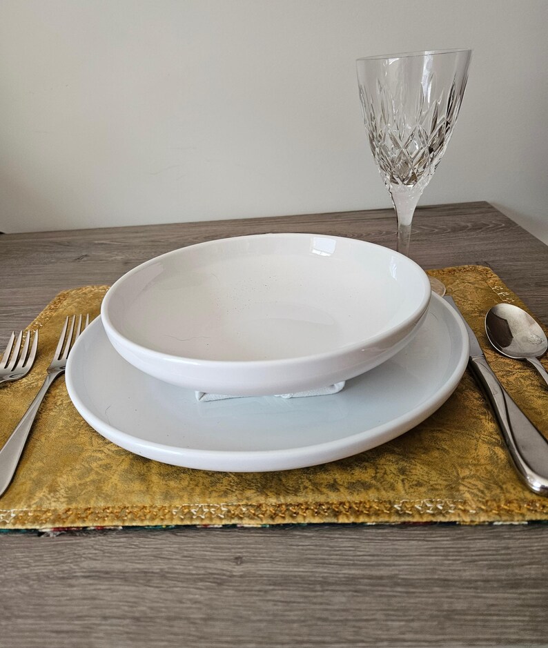 May include: A white ceramic bowl and plate set on a gold placemat, with silverware and a crystal wine glass. The bowl sits atop the plate, creating a layered effect. The setting is on a wooden table.