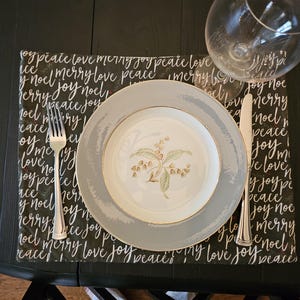 May include: A formal place setting on a black table. A white and gray plate with a floral design sits on a black placemat with white script. A fork and knife are on either side of the plate, and a wine glass is in the background.