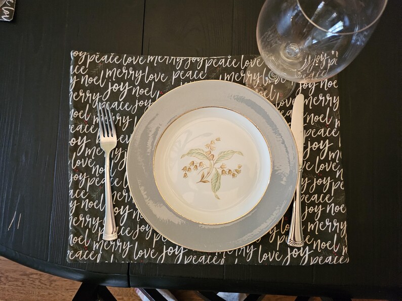 May include: A formal place setting on a black table. A white and gray plate with a floral design sits on a black placemat with white script. A fork and knife are on either side of the plate, and a wine glass is in the background.