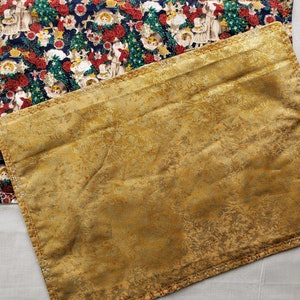 May include: Two rectangular placemats. One features a navy blue background with a Christmas-themed pattern of angels, Santa Claus, and red roses. The other is gold with a subtle floral design and a decorative stitched border.