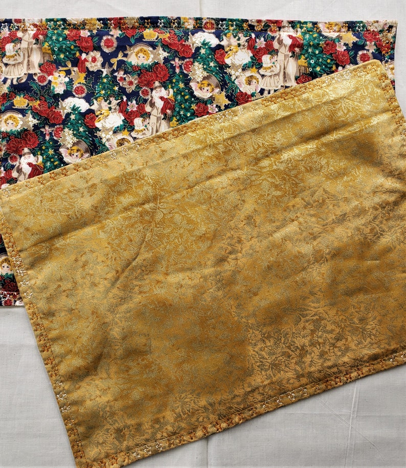 May include: Two rectangular placemats. One features a navy blue background with a Christmas-themed pattern of angels, Santa Claus, and red roses. The other is gold with a subtle floral design and a decorative stitched border.