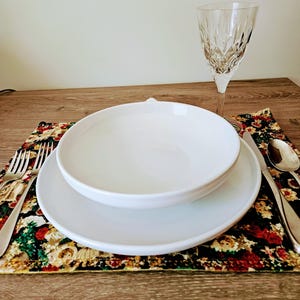 May include: A place setting with a white bowl and plate, a crystal wine glass, and silverware on a floral placemat. The placemat has a dark background with red, green, and gold floral patterns. The table setting is on a wooden surface.