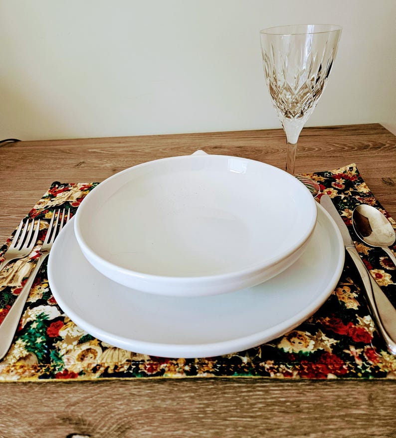 May include: A place setting with a white bowl and plate, a crystal wine glass, and silverware on a floral placemat. The placemat has a dark background with red, green, and gold floral patterns. The table setting is on a wooden surface.