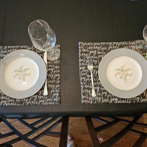 May include: A table setting with two plates, forks, knives, and wine glasses. The plates have a gray rim and a floral design. Placemats with the words "peace," "love," and "joy" are under the plates.