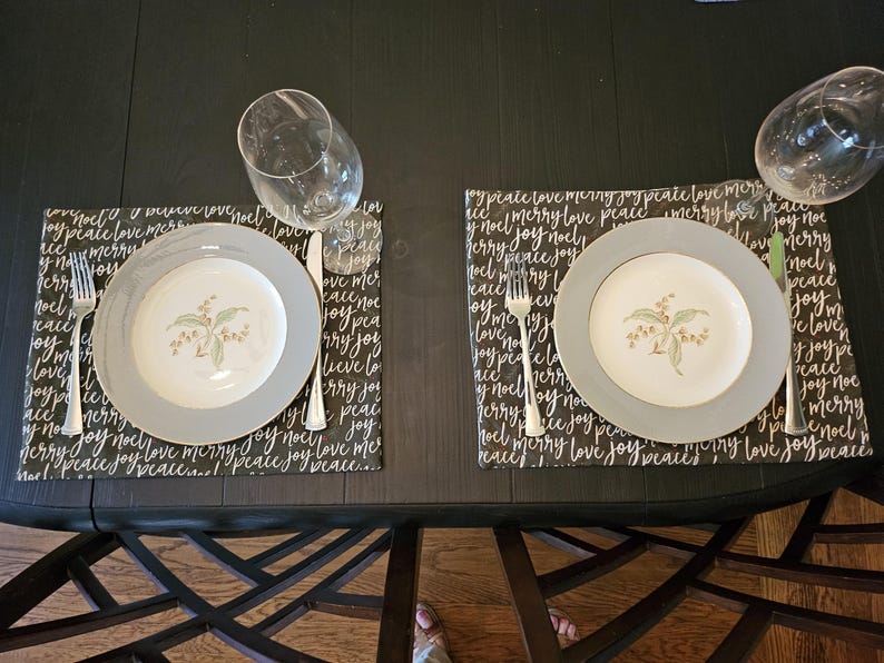 May include: A table setting with two plates, forks, knives, and wine glasses. The plates have a gray rim and a floral design. Placemats with the words "peace," "love," and "joy" are under the plates.