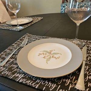 May include: A table setting with a gray-rimmed plate featuring a floral design, a fork, and a knife. The plate rests on a black placemat with white script that reads "joy, peace, love, merry, noel". A wine glass is also present.