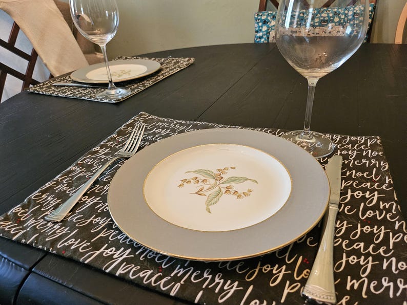 May include: A table setting with a gray-rimmed plate featuring a floral design, a fork, and a knife. The plate rests on a black placemat with white script that reads "joy, peace, love, merry, noel". A wine glass is also present.