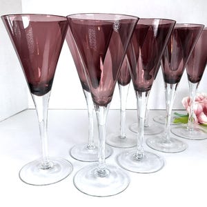 Puede incluir: Juego de seis copas de vino de vidrio transparente con un tinte morado oscuro. Las copas tienen un tallo alto y delgado y una copa ancha y acampanada.