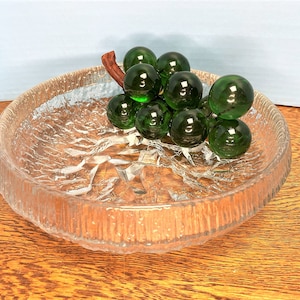 Puede incluir: Un cuenco de vidrio transparente con una superficie texturizada, que presenta un racimo de uvas de vidrio verde con un tallo marr&oacute;n.
