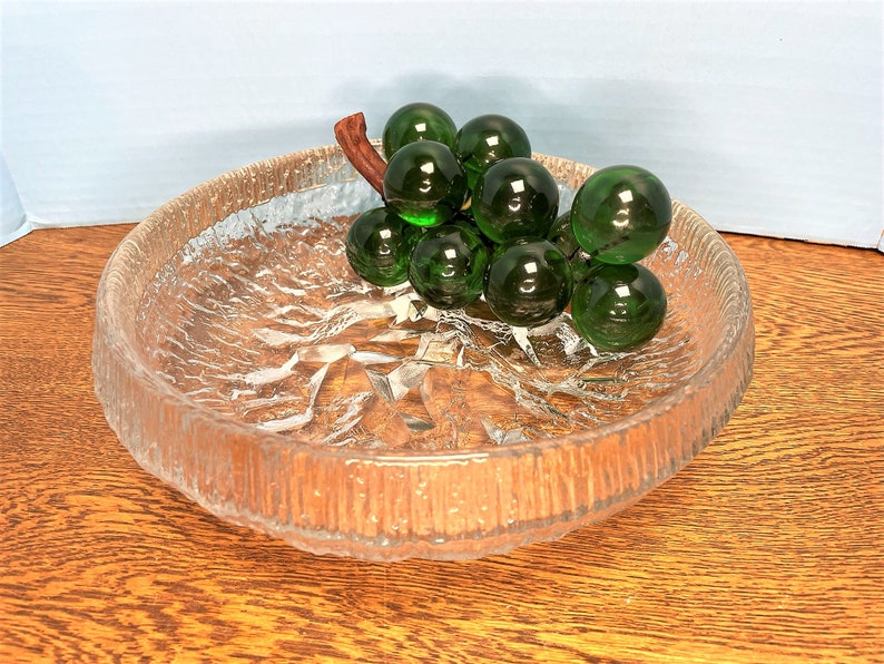 Puede incluir: Un cuenco de vidrio transparente con una superficie texturizada, que presenta un racimo de uvas de vidrio verde con un tallo marr&oacute;n.