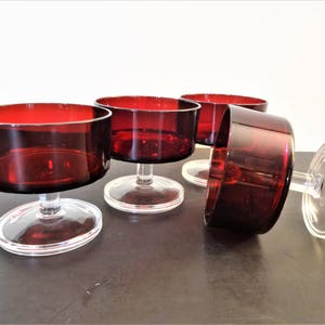 May include: Set of four vintage dessert glasses. Each glass features a clear, circular base and a stem supporting a deep red bowl. The glasses are arranged on a dark surface, with one glass tipped on its side.