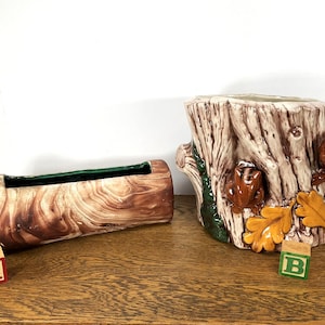 May include: Two ceramic planters shaped like tree stumps. The larger planter has a brown, white, and green glaze with a squirrel design. The smaller planter is brown and white with a green glaze inside. Both planters are sitting on a wooden surface.