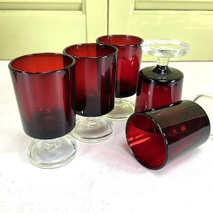 May include: A set of six ruby red stemmed glasses. The glasses have a clear base and a deep red bowl. One glass is lying on its side. The glasses are arranged on a white surface.