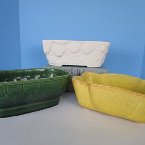 May include: Three ceramic planters in different colors and styles. The planter on the bottom is yellow with a speckled finish. The middle planter is green with a textured design. The top planter is white with a raised floral design.