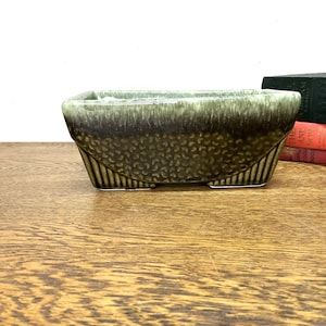 May include: A rectangular green and brown ceramic planter with a textured surface. The planter has four small feet and a slightly flared lip.