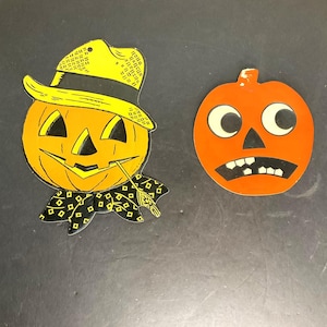 May include: Two vintage Halloween paper cutouts of jack-o'-lanterns. One is wearing a yellow hat and a black and white bow tie. The other has large eyes and a wide mouth.