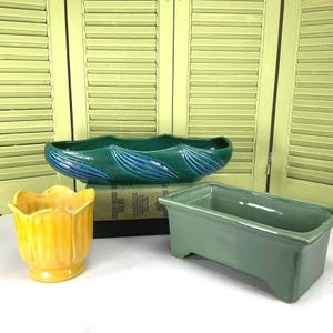 May include: A collection of ceramic planters in various shapes and colors. Includes a teal, oblong planter with blue accents, a yellow tulip-shaped planter, and a rectangular green planter. The planters are displayed on a white surface.