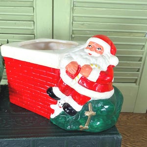 May include: A ceramic Christmas planter featuring Santa Claus. The planter is red and white, with Santa Claus in a red suit and hat, sitting on a green base. The planter is designed to look like a chimney.