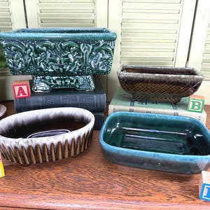May include: Four ceramic planters of varying shapes and colors. One is a rectangular dark green piece with ornate relief, one is a brown textured rectangular planter, one is an oval brown and white drip-glaze planter, and one is a teal rectangular dish.