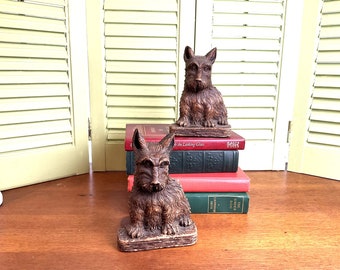May include: Two wooden bookends shaped like Scottish Terriers. The bookends are brown and have a realistic appearance. They are sitting on top of a stack of books.