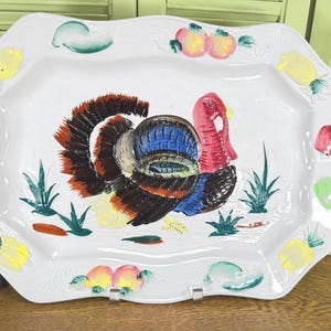 May include: A white rectangular ceramic serving platter with scalloped edges, featuring a central hand-painted turkey with brown, blue, and red plumage. The border is decorated with embossed and painted motifs of yellow pineapples and red fruits.