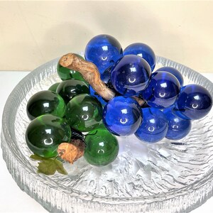 Puede incluir: Un racimo de uvas de vidrio con uvas azules y verdes en un plato de vidrio transparente con una superficie texturizada.
