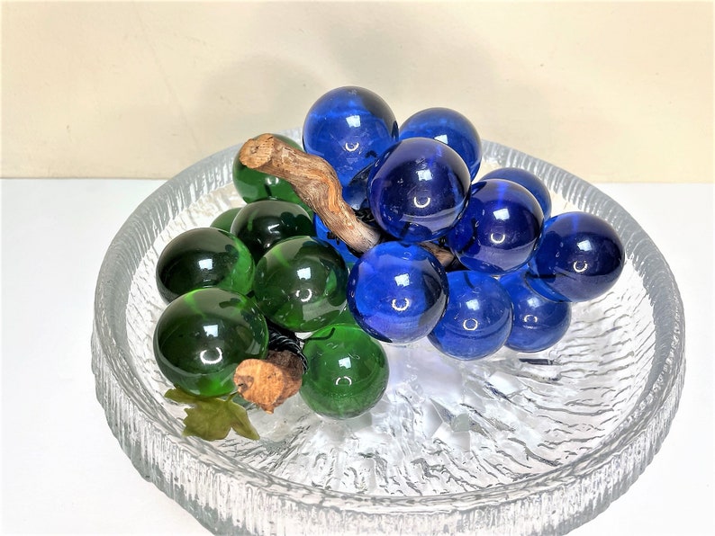 Puede incluir: Un racimo de uvas de vidrio con uvas azules y verdes en un plato de vidrio transparente con una superficie texturizada.
