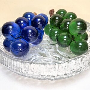 Puede incluir: Un cuenco de vidrio transparente con una superficie texturizada contiene dos racimos de uvas de vidrio. Un racimo es azul y el otro es verde.