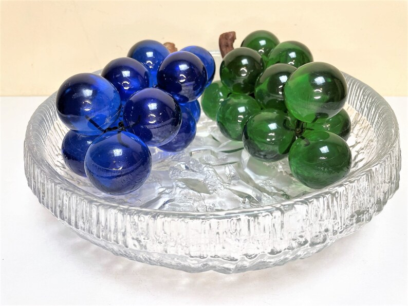 Puede incluir: Un cuenco de vidrio transparente con una superficie texturizada contiene dos racimos de uvas de vidrio. Un racimo es azul y el otro es verde.