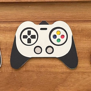 May include: Three white and black video game controller cutouts with colorful buttons and directional pads are arranged on a wooden surface. The controllers have a classic design, with a central directional pad and colored buttons.