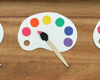 May include: Three white paint palettes with a paintbrush on each. The palettes have eight circular wells filled with different colours of paint. The colours are red, orange, yellow, green, blue, purple, pink, and brown.