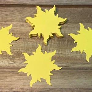 May include: Several yellow sun-shaped cutouts are arranged on a wooden surface. The suns have pointed rays and are in various positions, with one stack of cutouts. The background is a light brown wood grain.