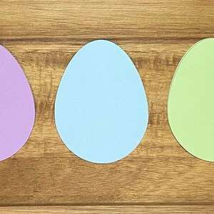 May include: Five pastel colored paper Easter eggs in a row on a wooden surface. The eggs are pink, purple, light blue, light green, and yellow.