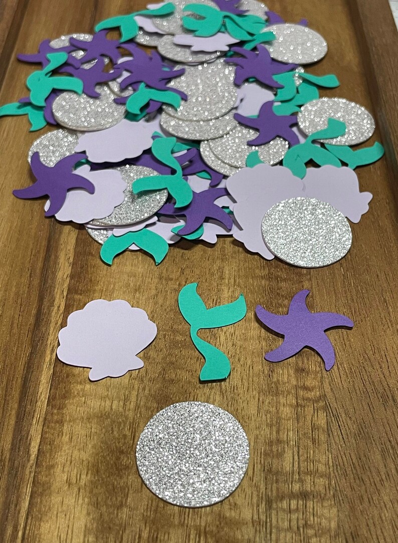 May include: A pile of purple, teal, and silver glitter paper cutouts of seashells, starfish, and mermaid tails.