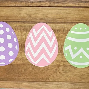 May include: Five paper Easter eggs in different colors and patterns. The eggs are yellow with white stripes, purple with white polka dots, pink with white zigzags, green with white swirls, and blue with white stripes.