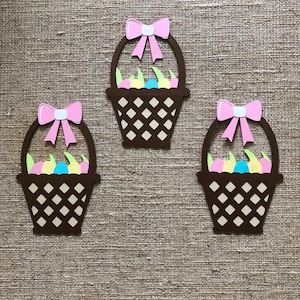 May include: Three brown paper Easter baskets with pink bows and colorful eggs inside. The baskets are cut out and have a checkered pattern.