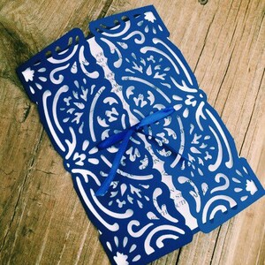 May include: A rectangular invitation card featuring a deep blue laser-cut paper overlay with intricate swirling patterns. The overlay folds inward to reveal a white inner card with visible black text. A thin blue ribbon is tied in a bow at the center.